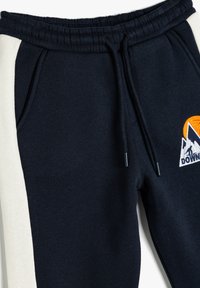 Marineblå sweatpants med cremefarvede sidelænger, snortræk i taljen og et broderet bjerglogo med orange og hvide accenter.