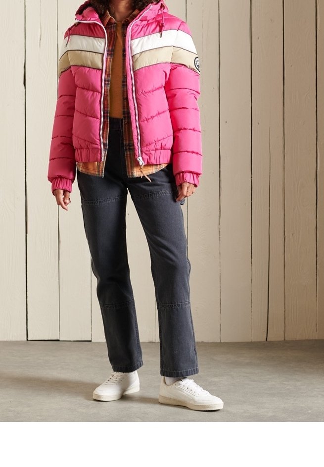 Lyserød puffer-jakke med hvide og beige striber, lynlås fortil og hætte. Båret over en ternet skjorte, kombineret med mørke jeans og hvide sneakers.