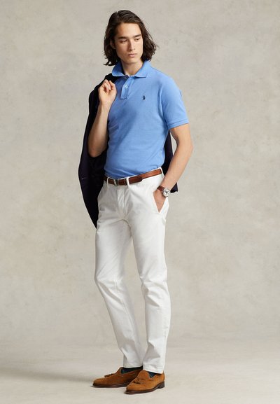 Jaunas vyras mėlynu polo marškinėliu, baltais kelnėmis, rudu diržu ir loafer batais, laikantis tamsiai mėlyną švarką ant peties, žiūrint į šoną prieš paprastą foną.