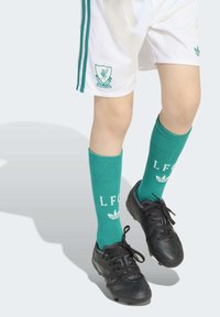 Zelene nogometne nogavice z belim napisom "LFC" in poudarki, nosene z črnimi športnimi copati in belimi kratkimi hlačami, ki imajo teal stranske črte in logo.