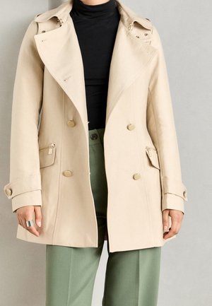 Manteau beige à double boutonnage avec des boutons dorés, porté sur un haut noir et un pantalon vert, avec une bague visible à la main gauche.