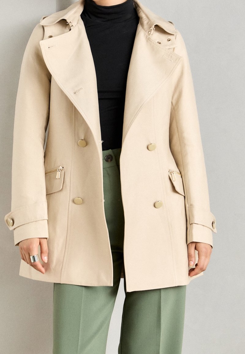 Manteau beige à double boutonnage avec des boutons dorés, porté sur un haut noir et un pantalon vert, avec une bague visible à la main gauche.