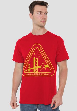Red cotton t-shirt featuring a yellow triangular graphic with text and illustrations of a bridge and starship motif. Comfortable fit.