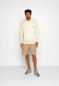 Lys beige hættetrøje med frontlomme og logo, parret med khaki shorts og grå træningssko. Neutrale farver, afslappet pasform.