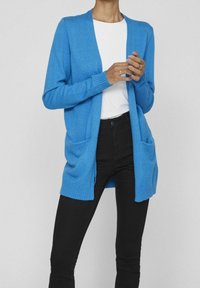 Helles blaues, gestricktes Cardigan mit offener Vorderseite, langen Ärmeln und zwei Fronttaschen. Über einem weißen Oberteil getragen und mit schwarzen Hosen kombiniert.