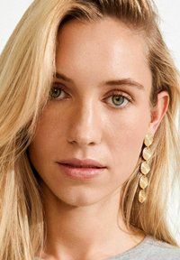 Kuldne spiraalkujulised kõrvarõngad tekstuurse viimistlusega, langevad alla lineaarse mustriga, kandmas valge juuksevärviga ja heleda nahaga modell.