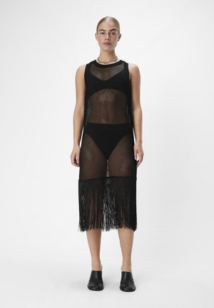 Svart strikket cover-up kjole med en tettsittende bodice, frynsehem og gjennomsiktige deler. Båret over matchende svart bikiniunderdel.
