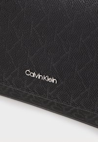 Crna torba s teksturom i geometrijskim uzorkom. Sadrži srebrni logo Calvin Klein na prednjoj strani. Glatka površina s izraženim linijama i oblicima.