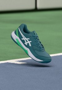 Türkisfarbener Sportschuh mit Mesh-Obermaterial, weißen Akzenten und grünen Details. Für den Sport konzipiert, mit Dämpfung und einer strukturierten Sohle.