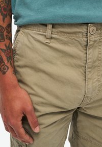 Beige cargoshorts met een gestructureerd oppervlak, voorzakken en een knoopsluiting. Draagt een blauwe crewneck t-shirt dat zichtbaar is bovenop.