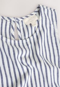 Bílá lněná látka s vertikálními námořnicky modrými pruhy ukazující kulatý výstřih a štítek na oblečení s nápisem "Contains linen Next."