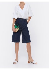 Nainen, jolla on päällään valkoinen V-pääntieblossi, tummat farkkishortsit polven ylle, valkoiset korot sandaalit, ja hän pitää pientä pyöreää olkipussukkaa, jossa on vihreät kahvat.