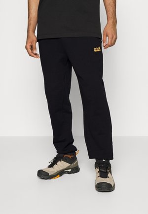 Person iført sorte Jack Wolfskin-bukser og beige vandresko, stående på en lysegrå gulv mod en hvid væg.