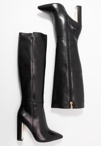 Svarta läderknähöga stövlar med spetsiga tår och blockklackar. Har en guldzipperdetalj och en slät, glänsande yta.