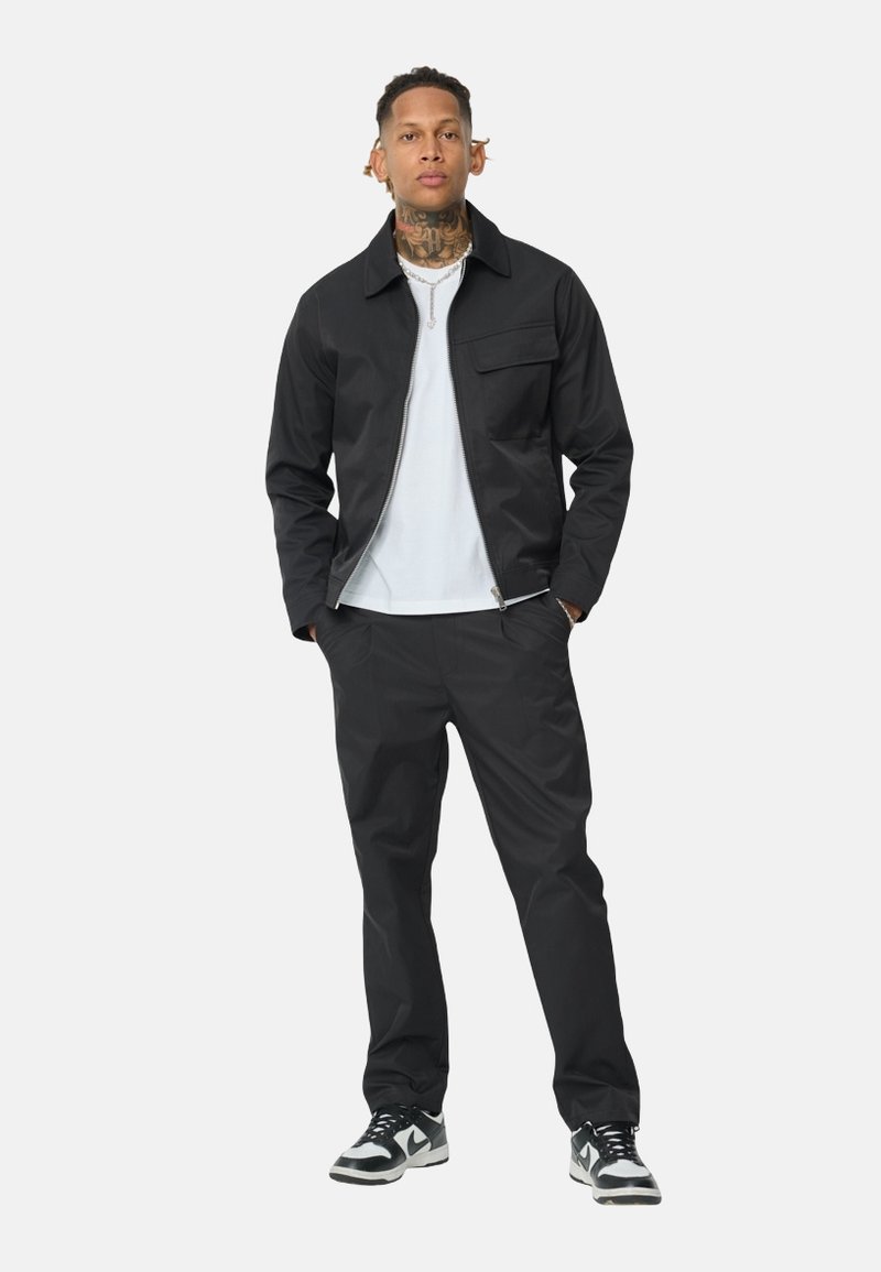 Veste noire légère avec col, fermeture éclair à l'avant, poche unique. Associée à un pantalon noir assorti et des baskets noir et blanc.