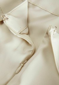 Beige stof met een gladde textuur, voorzien van een ritssluiting en twee zichtbare ritsen. Minimalistisch ontwerp met nette stiksels.