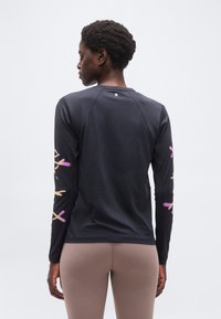 Άτομο με την πλάτη προς τα εμπρός που φοράει μαύρο πουκάμισο μακρυμάνικο με ροζ και κίτρινα σχέδια στα μανίκια και μπεζ παντελόνι, σε απλό φόντο.