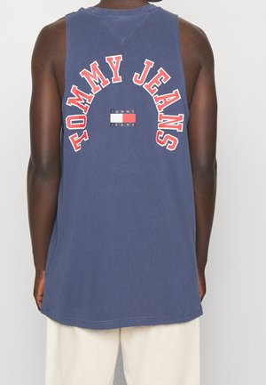 Personne portant un haut sans manches bleu marine avec le texte « TOMMY JEANS » et un petit logo rouge, blanc et bleu dans le dos, associé à un pantalon blanc.