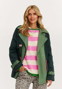 Zelená nepromokavá bunda s dvojitým zapínáním, nošená přes krémový svetr s růžovými pruhy a kalhoty s leopardím vzorem.