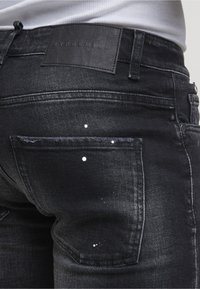 Jean en denim noir avec des éclaboussures de peinture blanche sur la poche arrière et une étiquette de marque en cuir à la taille.