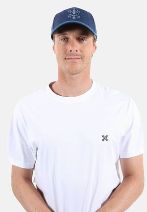 Homme portant un T-shirt blanc uni avec un petit logo sur la poche poitrine et une casquette de baseball bleue avec des motifs blancs.