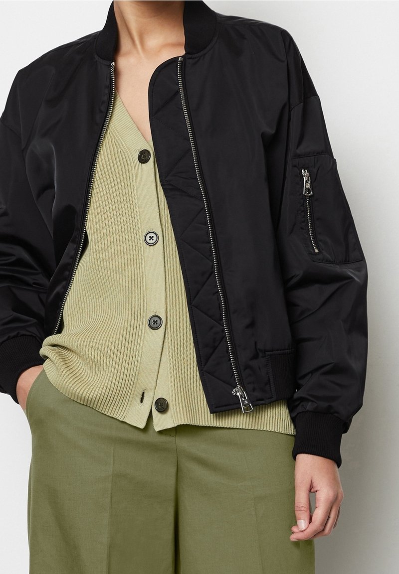 Chaqueta bomber negra con puños acanalados, cremallera frontal y tela texturizada. Llevada sobre un cárdigan verde claro acanalado y pantalones verde oliva.