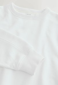 Hvid langærmet sweatshirt med ribstrikket rund hals og manchetter, lagt fladt ud på en hvid overflade, der viser en del af ærmet og kraven.