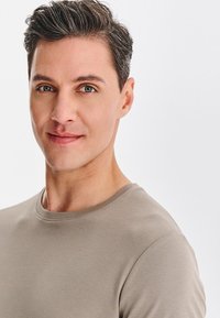 Mężczyzna z krótkimi włosami, ubrany w jasnobrązową, koszulę z krótkim rękawem i okrągłym dekoltem. Gładka skóra i neutralny wyraz twarzy na białym tle.