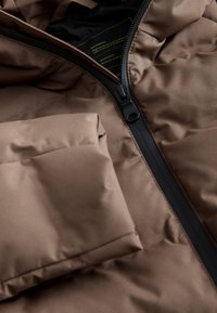 Pruun puffer-jakk, millel must tõmblukk ja rinnatasku, valmistatud tekstuurse, veekindla materjali ja kirjuga täidisega.