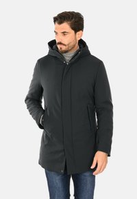 Cappotto nero isolante con cappuccio, dotato di chiusura con zip e bottoni a pressione, tasche laterali e finitura liscia e opaca. Indossato sopra a un maglione leggero.