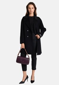 Cappotto doppiopetto in lana nera, top aderente nero, pantaloni affusolati e décolleté nere a punta. Porta una borsa in pelle bordeaux con tracolla a catena.