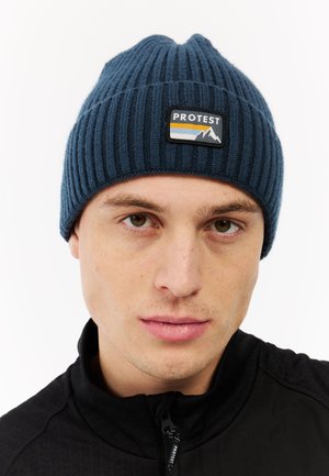 Knit blue beanie with ribbed texture, featuring a black patch with "PROTEST" and mountain graphic. Worn with a zip-up black jacket.