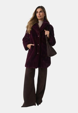 Femme portant un manteau en fourrure violet foncé, un pantalon large marron, tenant un sac à main marron, debout devant un fond clair uni.
