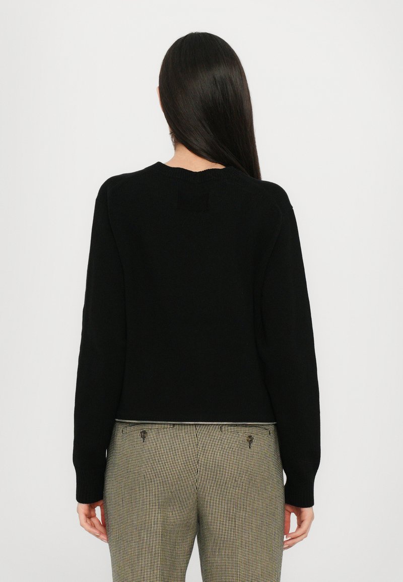 Black knitted sweater with a rounded neckline and long sleeves. The back features a small patch detail. Worn with checkered trousers.