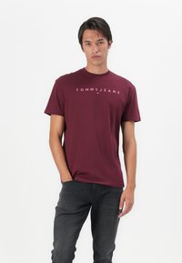 Maronnes Baumwoll-T-Shirt mit Rundhalsausschnitt, mit dem weißen "TOMMY JEANS"-Logo auf der Brust. Getragen mit dunkelgrauen Jeans.