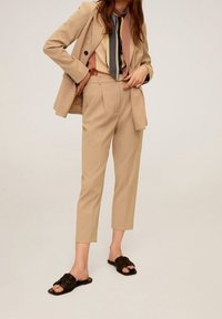Abito beige su misura con blazer doppiopetto e pantaloni cropped a vita alta. Include una blusa a righe multicolore e sandali neri a ciabatta.