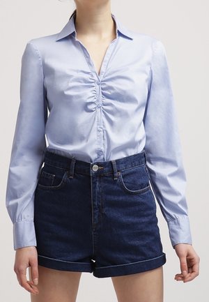 Vrouw die een lichtblauw geplooid blouse met knopen draagt, ingestopt in een high-waisted donker denim short met opgerolde zoom, staand tegen een effen achtergrond.