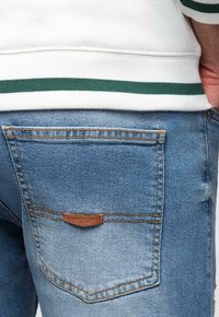 Jean en denim bleu clair avec une étiquette en cuir sur la poche arrière, présentant des coutures contrastantes et une coupe classique, associé à un sweat-shirt blanc.