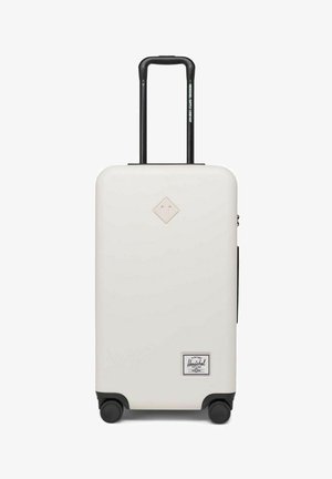 Valise rigide blanche avec une poignée rétractable noire, quatre roues noires et un patch avec le logo de la marque en bas à droite.