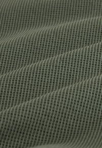 Tekstiili tummanvihreänä, hienolla ruutukuvioinnilla. Materiaali näyttää pehmeältä, sillä on sileä pinta ja hillitty kiilto valon leikin vuoksi.