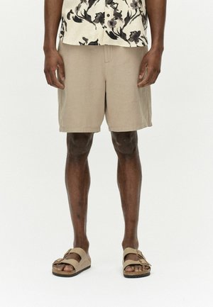 Beige Leinen-Shorts mit Seitentaschen, elastischem Bund und entspanntem Schnitt, kombiniert mit braunen Sandalen, die zwei breite Riemen haben.