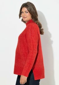 Heller roter Pullover mit hohem Kragen, langen Ärmeln und Seitenschlitzen. Weiche Textur mit gerippten Bündchen und Saum. Über dunklen Jeans getragen.