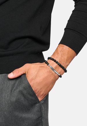 Männliche Hand in der grauen Hosentasche mit zwei schwarzen Perlenarmbändern, eines mit einer horizontalen silbernen Platte, vor einem schwarzen Langarmshirt.