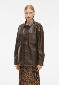 Veste en cuir marron avec col, deux poches avant et fermeture à boutons-pression, portée sur une longue robe marron à motifs.
