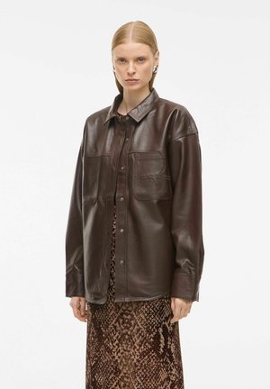 Veste en cuir marron avec col, deux poches avant et fermeture à boutons-pression, portée sur une longue robe marron à motifs.