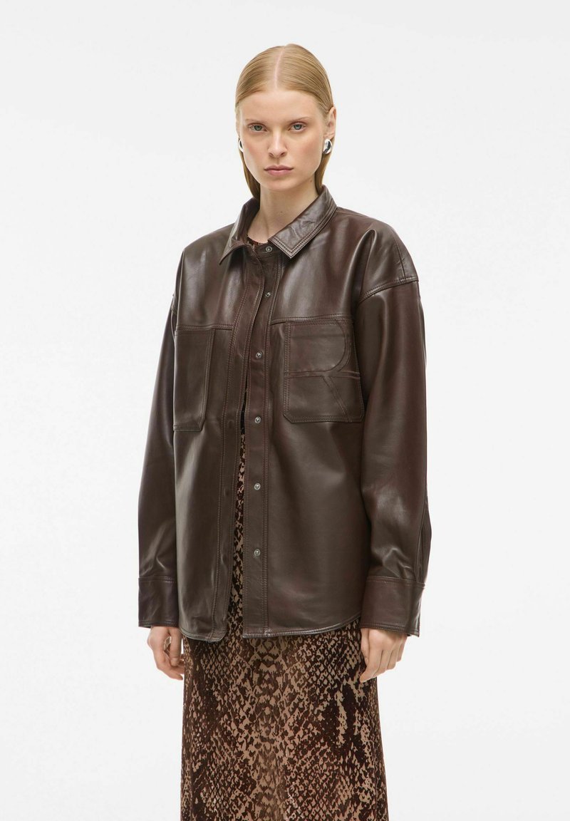 Veste en cuir marron avec col, deux poches avant et fermeture à boutons-pression, portée sur une longue robe marron à motifs.