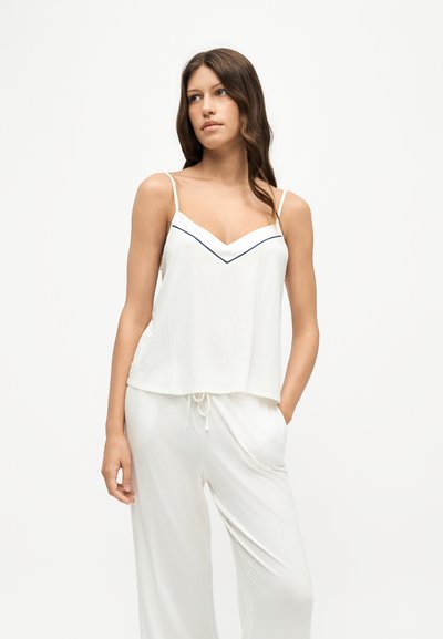 Femme aux longs cheveux bruns portant un haut blanc sans manches avec de fines bretelles et un pantalon blanc ample assorti, debout devant un fond blanc uni.