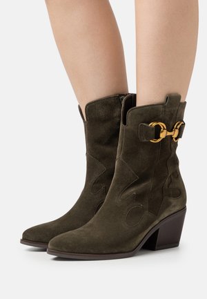 Bottes brunes mi-mollet en daim avec talons blocs, couture décorative et détail de boucle en métal doré, portées sur des jambes nues.