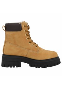 Stivaletto in suede color beige con suola in gomma nera, occhielli dorati, dettagli marroni e punta arrotondata. Presenta un laccio frontale e colletto imbottito.