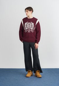 Μαρόν πλεκτό πουλόβερ με λευκές λεπτομέρειες και λογότυπο Adidas, συνδυασμένο με μαύρα παντελόνια σε φαρδιά γραμμή και καφέ μπότες. Απλός, ανεπίσημος σχεδιασμός.